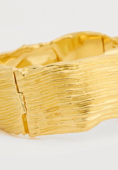 Φαρδύ χρυσό βραχιόλι με υφή, άνισες οριζόντιες ραβδώσεις και κούμπωμα ορατό στη μία πλευρά, εμφανίζεται σε απλό ανοιχτό φόντο.