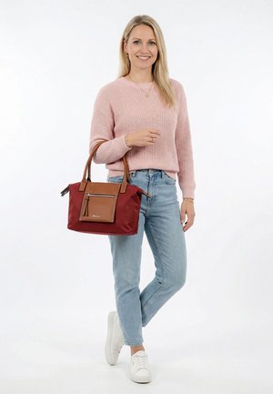 Femme portant un pull rose, un jean bleu clair et des baskets blanches, tenant un sac à main rouge et marron, debout sur un fond blanc.