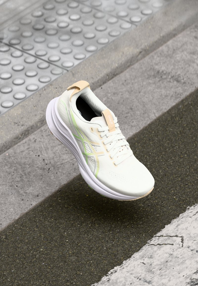 Zapato de correr blanco con parte superior texturizada, detalles en verde claro y una lengüeta en el talón color beige. Cuenta con una suela acolchada y cierre de cordones.