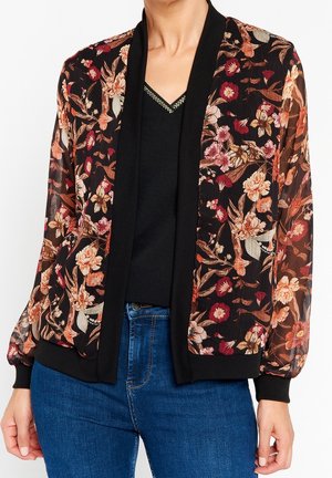 Sort gennemsigtig blomster bomberjakke med ribbet krave og manchetter, båret over en sort V-hals top og blå jeans.