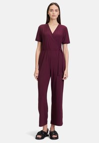 Bordeauxkleurige jumpsuit in wikkelstijl met korte mouwen, strik in de taille en een wijde pijpenstijl. G gedragen met zwarte geknoopte sandalen.