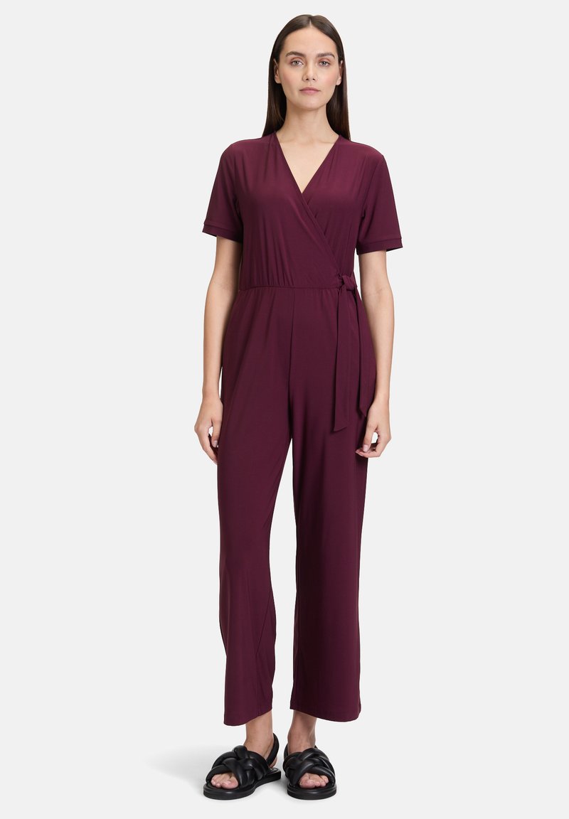 Bordeauxkleurige jumpsuit in wikkelstijl met korte mouwen, strik in de taille en een wijde pijpenstijl. G gedragen met zwarte geknoopte sandalen.