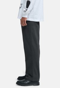 Zijaanzicht van een persoon die zwarte rechte broek, zwarte leren schoenen en een wit shirt met lange mouwen draagt, met zwarte grafische prints op de mouw.