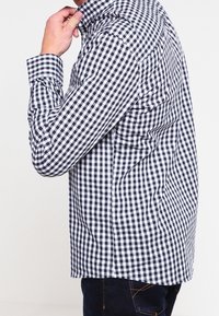 Uomo che aggiusta il colletto di una camicia a maniche lunghe bianco e nero a scacchi, indossando jeans blu scuro, in piedi contro uno sfondo uniforme.