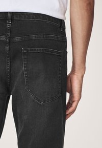 Czarne dżinsy z denimu o dopasowanym kroju, z pojedynczą kieszenią z tyłu i subtelnymi detalami szwów. Materiał wydaje się lekko fakturowany.