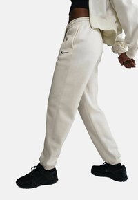 Beige Sweatpants mit elastischem Bund, seitlichen Taschen und einem kleinen schwarzen Nike-Logo am linken Oberschenkel, kombiniert mit schwarzen Sportschuhen.