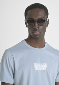 Camiseta azul claro hecha de tela suave con cuello redondo, con un diseño de salpicaduras blancas y un logo en el pecho; modelo con gafas de sol oscuras.