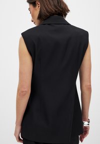 Blazer sin mangas negro con hombros estructurados, cuello afilado y corte tailored. Tela suave y detalle de costura mínima en la parte posterior.