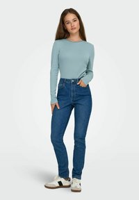 Lys blå genser med lange ermer kombinert med høytlivs mørkeblå jeans og hvite joggesko med sorte detaljer, med en tettsittende passform og glatt tekstur.