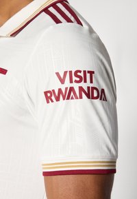 Camisola de desporto branca com realces em castanho-avermelhado, padrão texturizado e logótipo "Visit Rwanda" na manga, com riscas amarelas e castanho-avermelhadas na gola.