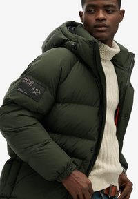 Zielona puchowa kurtka z kapturem, z zapięciem na zamek, z dzianinowym swetrem pod spodem i naszywką na lewym rękawie.