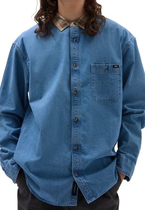 Persoon draagt een blauw denim overhemd met knopen, een beige geruite kraag, een borstzak en heeft handen in de zakken van donkere broeken.