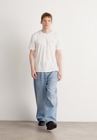 Hvit kortermet T-skjorte med en liten logo, kombinert med lyseblå løstsittende jeans og svarte sko. Nøytral bakgrunn med minimal tekstur.