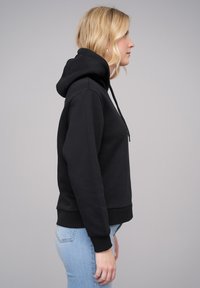 Black hoodie made of soft fabric, fitted design, kangaroo pocket, adjustable drawstring hood, ribbed cuffs and hem. Profile view.