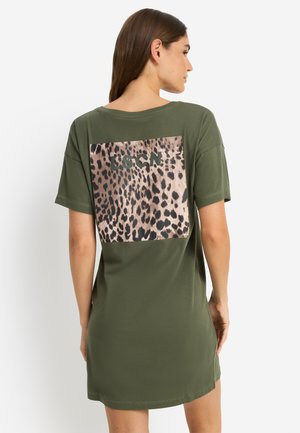 Sieviete, kas valkā olīvzaļas krāsas īsas piedurknes kleitu ar lielu leoparda raksta kvadrātu un "LSCN" tekstu uz muguras, stāvot pret baltu fonu.