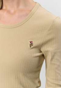 Khaki bordázott felső kerek nyakkivágással és hímezett díszítő folttal a bal mellen. Puha, testhez simuló anyag.