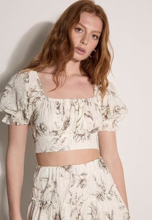 Junge Frau mit langen roten Haaren trägt ein weißes florales Eyelet-Croptop mit Puffärmeln und passendem hoch tailliertem Rock vor schlichtem Hintergrund.