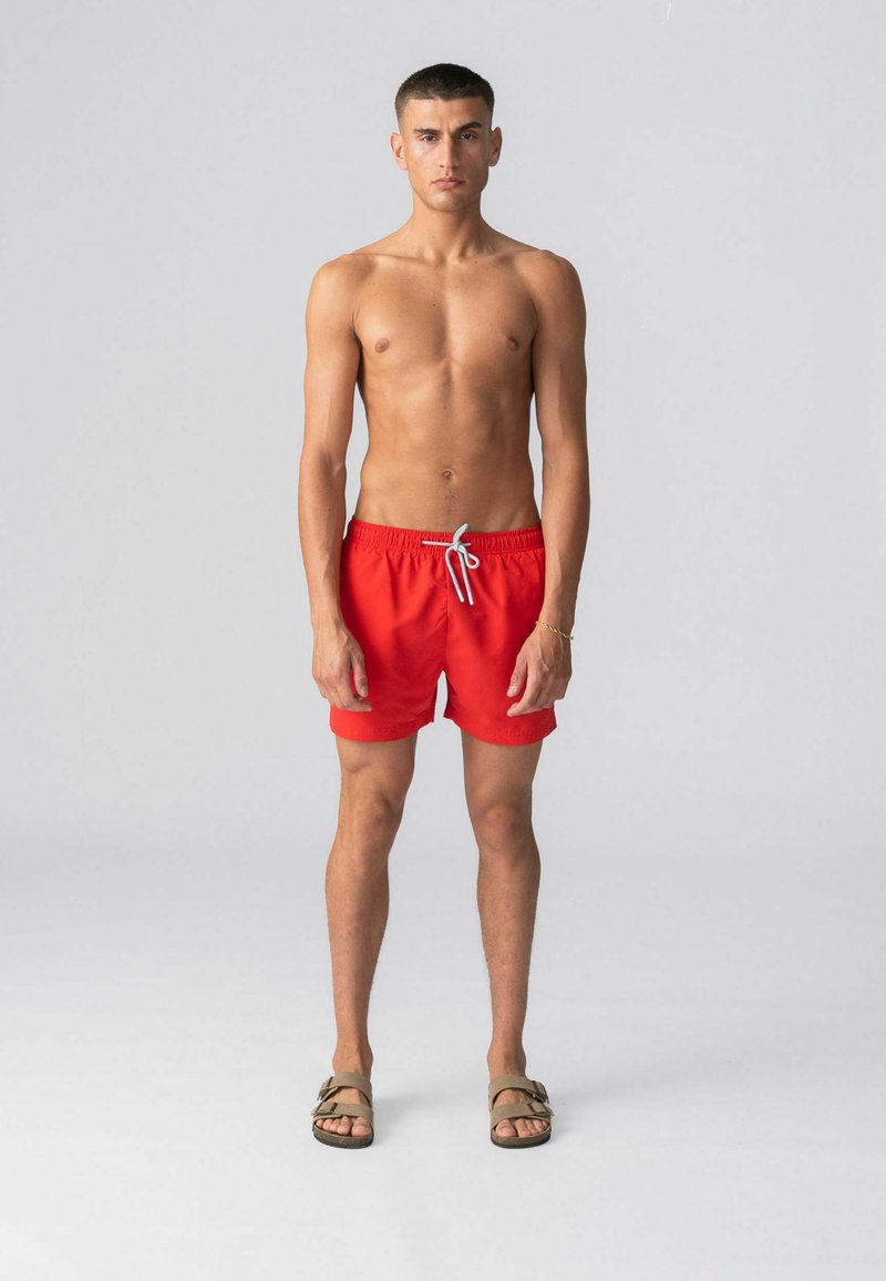 Fiatal férfi áll mezítláb, piros fürdőnadrágban és bézs szandálban, egyszínű világosszürke háttér előtt.