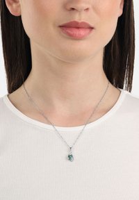 Collana d'argento con catena attorcigliata e pendente a goccia verde, con un piccolo accento trasparente. Indossata su una maglietta a costine leggera.