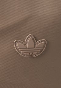 Fondo textil marrón con un logo de goma beige de Adidas Trefoil que presenta un diseño de tres hojas y tres líneas horizontales en el centro.