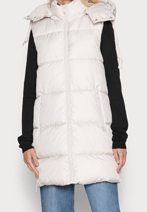 Kobieta ubrana w jasno-beżową pikowaną bezrękawnik z suwakiem z przodu i zapinanymi na suwak kieszeniami, nosząca pod spodem czarną bluzkę z długimi rękawami oraz niebieskie dżinsy.