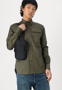 Zwarte schoudertas met twee ritssluitingen, gedragen over een olijfgroene blouse met knoopjes. De blouse heeft een borstzak en knopen.