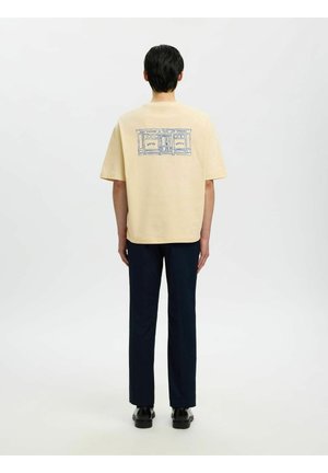 Osoba stoi tyłem, ubrana w beżową koszulkę z niebieskim szkicem witryny sklepowej oraz czarne spodnie na jednolitym białym tle.