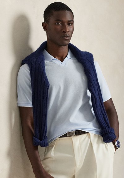 Marineblå strikket vest over en lyseblå piké, kombinert med kremfargede bukser. Har flat krage og teksturert strikkemønster.