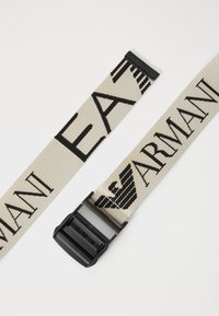Beige stoffen riem met zwart bedrukte tekst "EA" en "ARMANI", voorzien van een zwarte plastic gesp en een strak, plat ontwerp.