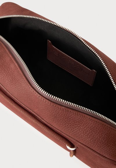 Sac en cuir marron avec une fermeture éclair argentée partiellement ouverte montrant une doublure intérieure noire et une étiquette marron cousue à l'intérieur.