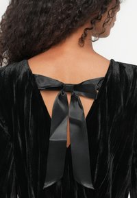 Musta samettinen mekko, jossa on syvä selkäaukko, sidottu satiininauhaan. Kankaan tekstuuri laskeutuu tyylikkäästi, korostaen kaula-aukon yksityiskohtia.