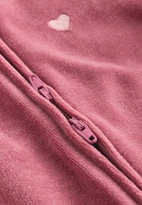 Chaqueta de forro polar en rosa con una textura suave. Presenta un pequeño corazón bordado y una cremallera rosa a juego en la parte delantera.