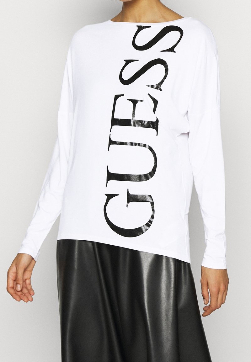 Frau, die ein weißes Langarmshirt mit großen vertikalen "GUESS"-Schriftzug in Schwarz und einen schwarzen Lederrock trägt.