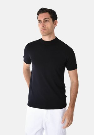 Giovane uomo con capelli scuri che indossa una maglietta nera aderente a maniche corte e pantaloni bianchi, in piedi con le mani dietro la schiena su uno sfondo semplice.