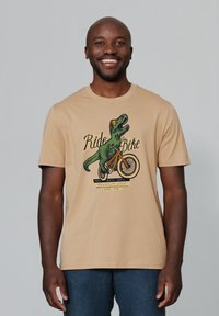 Beige Baumwoll-T-Shirt mit einem grünen Dinosaurier, der Fahrrad f�ährt, mit dem grafischen Text "Ride Bike" und zusätzlichen bunten Akzenten darunter.