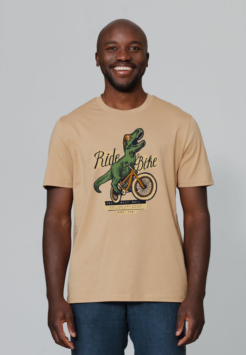 Beige Baumwoll-T-Shirt mit einem grünen Dinosaurier, der Fahrrad fährt, mit dem grafischen Text "Ride Bike" und zusätzlichen bunten Akzenten darunter.