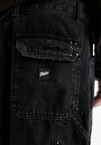 Acercamiento a un bolsillo de pantalones cargo de mezclilla negra con pequeñas manchas de pintura blanca y una etiqueta negra con logotipo blanco en la solapa del bolsillo.