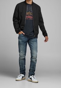 Svart bomberjacka över en marinblå t-shirt med tryck, kombinerad med ljusblå slitna jeans och vita sneakers med svarta detaljer.