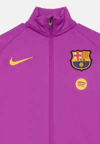 Lila zip-up sportjacka med gul Nike swoosh, FC Barcelona-emblem och gult Spotify-logotyp på framsidan av bröstet.