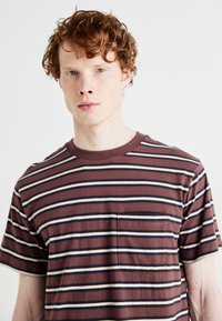 Brown short-sleeve t-shirt with horizontal stripes in beige and navy. Features a chest pocket and a crew neckline. Soft texture.