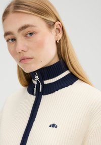 Sweter z dzianiny z pół zamkiem na kremowym tle, z wysokim kołnierzem w granatowe paski. Posiada zapięcie na zamek oraz małe haftowane logo na piersi.