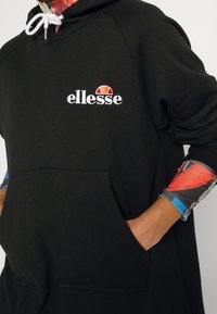 Personne portant un sweat à capuche ellesse noir avec des cordons blancs et des manches colorées avec des motifs abstraits rouges, bleus et gris.