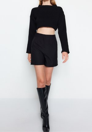 Frau trägt einen schwarzen, gerippten, bauchfreien Pullover, schwarze Shorts mit hoher Taille und kniehohe schwarze Lederstiefel vor weißem Hintergrund.