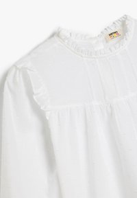 Witte blouse met lange mouwen, rimpelkragen, gestructureerde stof en een subtiel stippenpatroon. Heeft gathered details op de schouders en het lijfje.