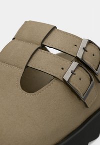 Gros plan d'une chaussure en daim beige avec deux sangles réglables et des boucles carrées argentées sur une semelle noire.