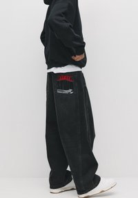 Sort denim bukser med vide ben med røde broderede flammer og logodetalje på baglommen; kombineret med en sort hættetrøje og hvide sneakers.