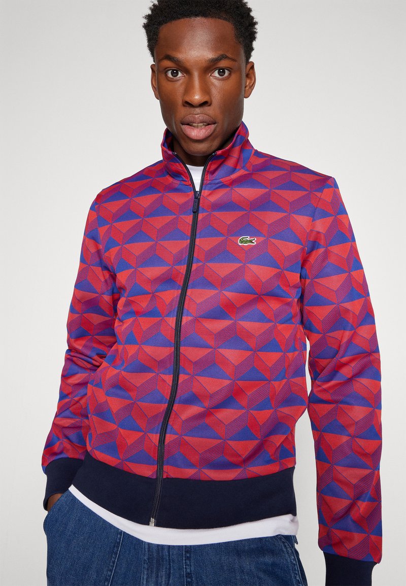 Veste zippée à motif géométrique rouge et bleu avec un col haut et un ourlet en rib à rayures navy. Présente un petit logo Lacoste sur la poitrine.
