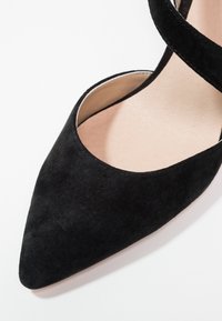 Chaussure plate en daim noir à bout pointu, avec une fine bride à la cheville et une semelle intérieure beige.