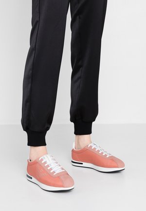 Pies con zapatillas bajas color rosa coral con cordones blancos y suelas en blanco y negro, combinadas con pantalones negros con puños remangados.