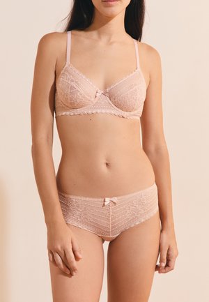 Femme portant un soutien-gorge et une culotte en dentelle rose clair assortis avec de petits nœuds, debout devant un fond beige uni.